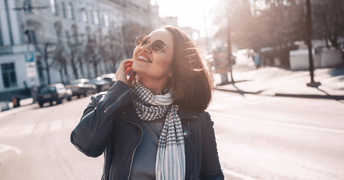 A happy, confident woman in sunglasses and a leather jacket smiling in the city, representing renewed vitality and wellness after hormone optimization and vaginal rejuvenation treatments.
