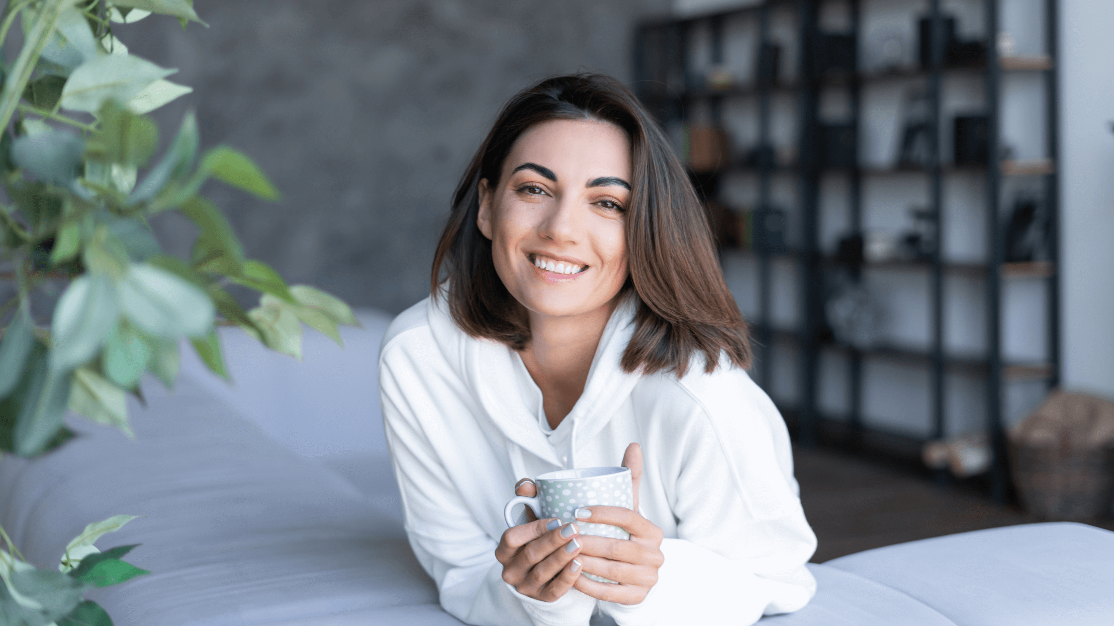 A smiling, relaxed woman in a white hoodie sitting on a couch and holding a coffee mug, representing the comfortable recovery and confidence achieved after a Morpheus8V or Votiva feminine rejuvenation treatment.