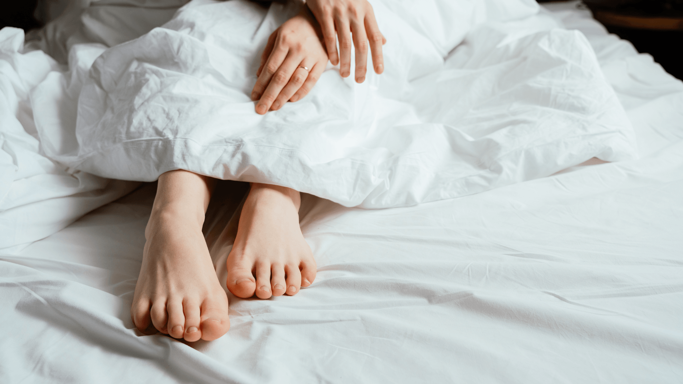 A woman’s feet resting under white bed linens, symbolizing the personal and intimate nature of discussing low libido and sexual wellness for women over 40.